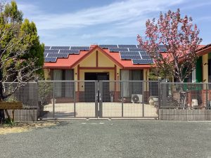 赤い屋根の施設建物外観。太陽光パネル搭載、フェンス付き。