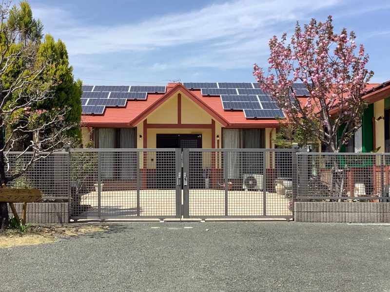 赤い屋根と太陽光パネルを備えた介護施設の建物外観。春の花が咲く庭が見える。